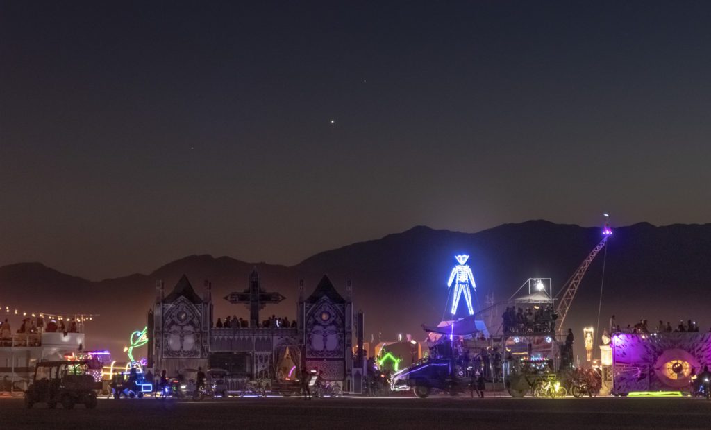 The Man at Burning Man with a bunch of art cars including a cathedral